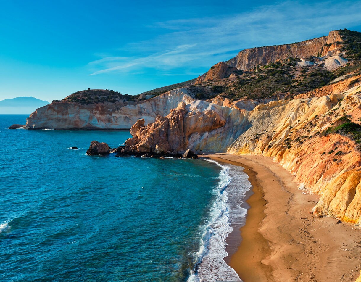 Agios Ioannis Beach with golden sand, turquoise waves and steep rocky cliffs in warm tones under a clear blue sky.