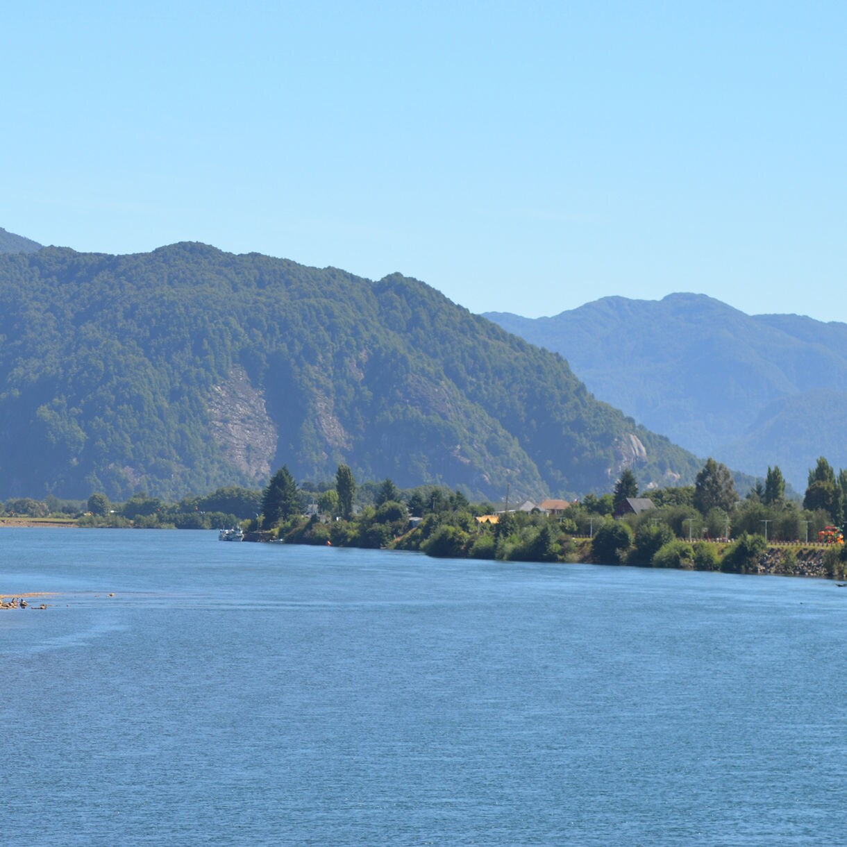 Wide river bordered by green trees with rolling forested mountains in the background under a clear blue sky.