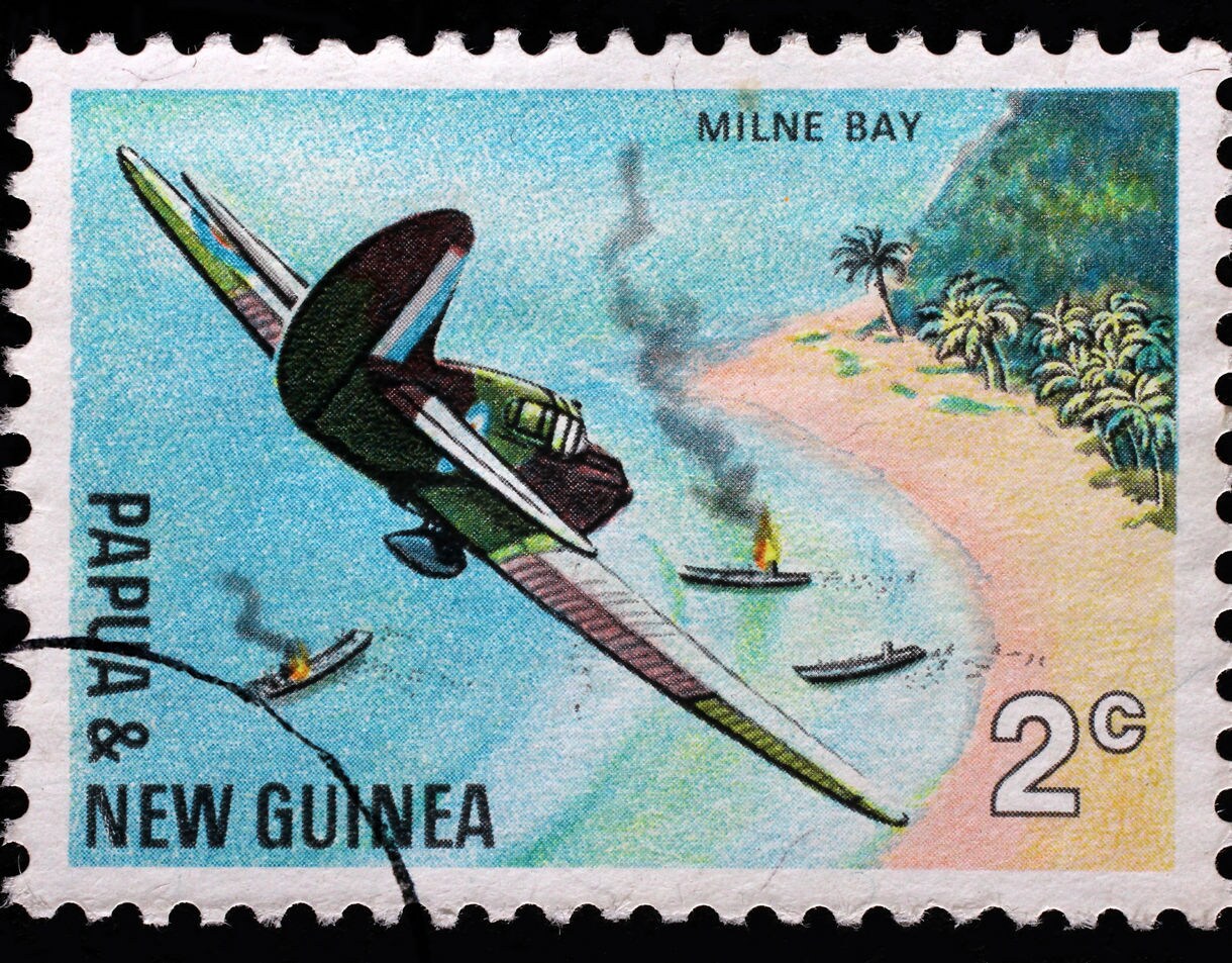 Vintage Papua and New Guinea postage stamp showing a military aircraft swooping over a tropical shoreline with several boats below, two of them burning and trailing smoke near a palm-lined beach.
