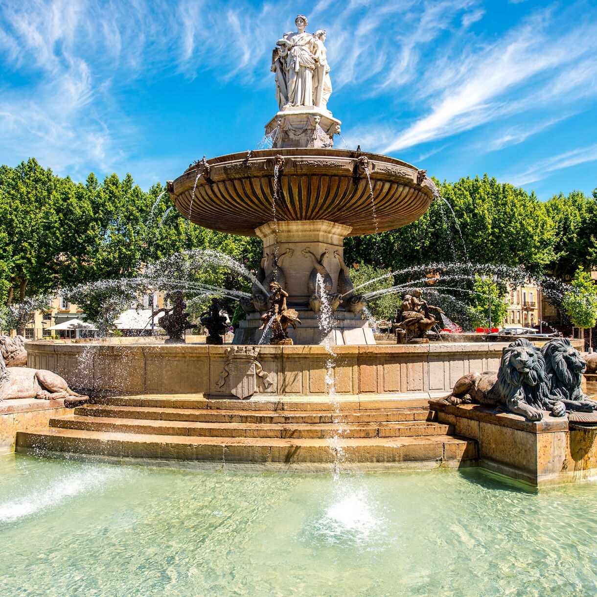 Ornate fountain in Aix-en-Provence featuring statues, lion sculptures and flowing water, set against lush green trees and a bright blue sky.