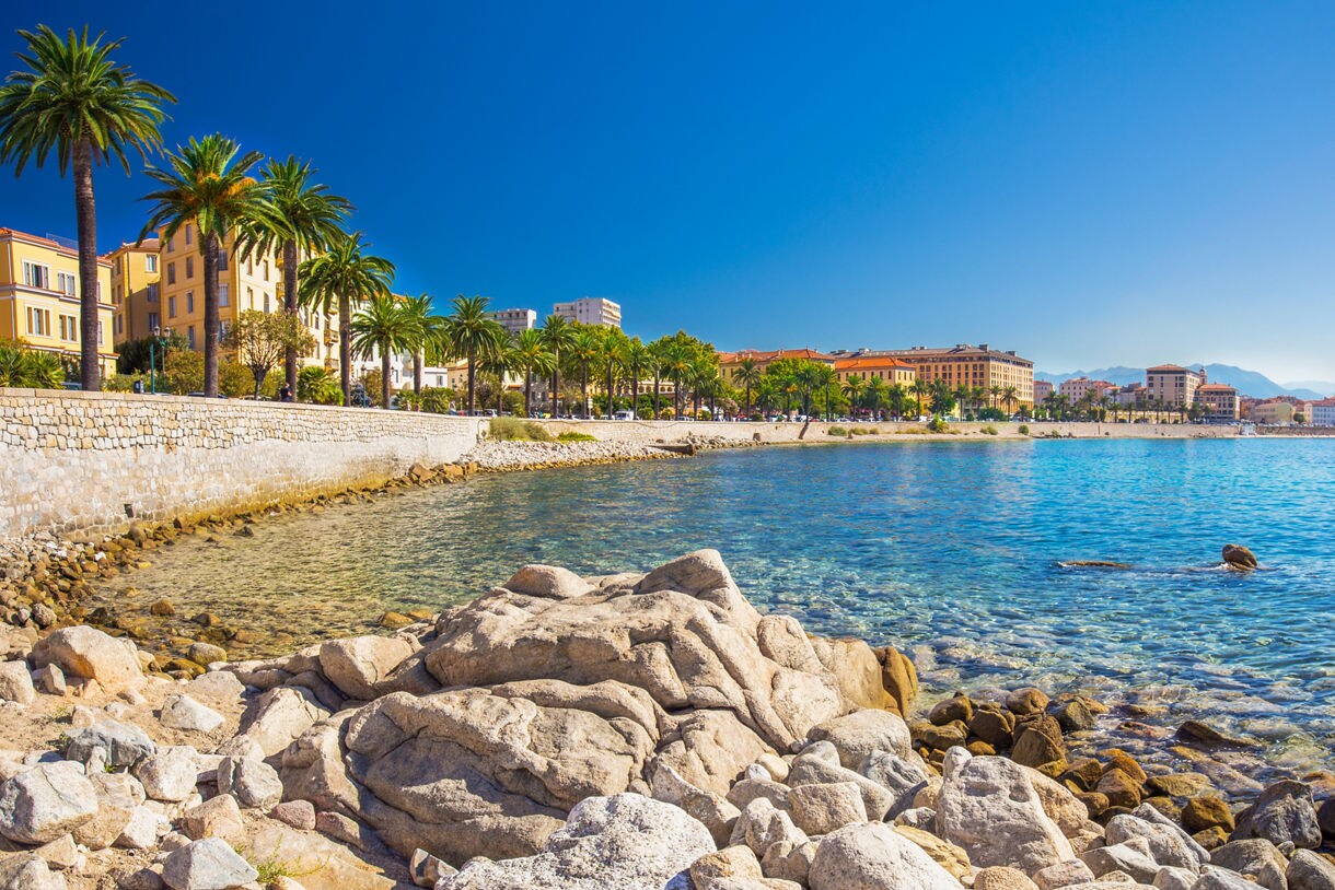 Coastal view of Ajaccio, France with palm trees, pastel buildings and clear blue water under a bright sky.
