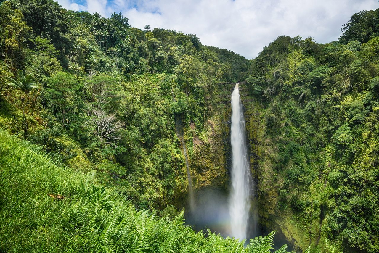 Tall waterfall dropping into a round basin, surrounded by steep green cliffs and dense tropical forest under a partly cloudy blue sky.