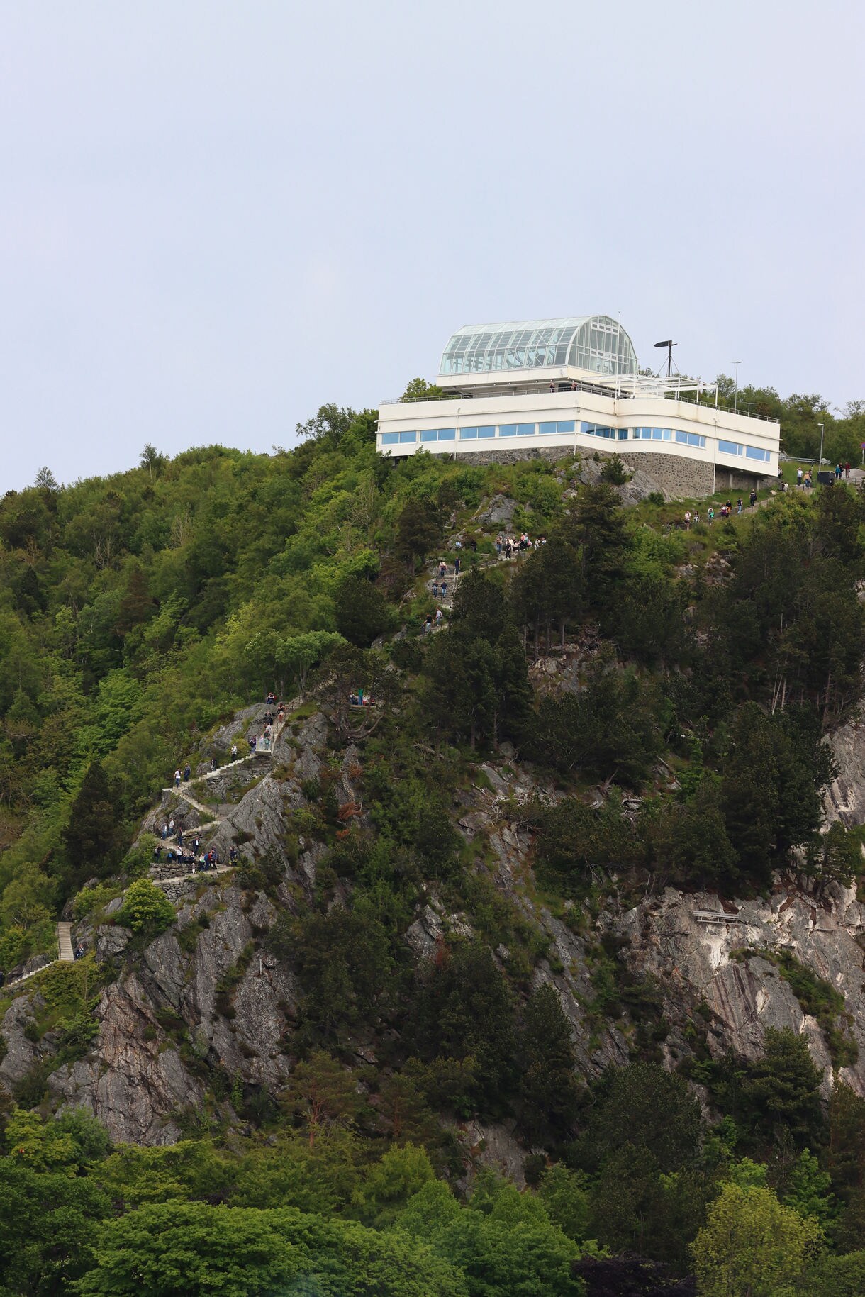 Modern glass-roofed Aksla Viewpoint building perched on a forested hillside above Ålesund, with a steep staircase trail visible below.