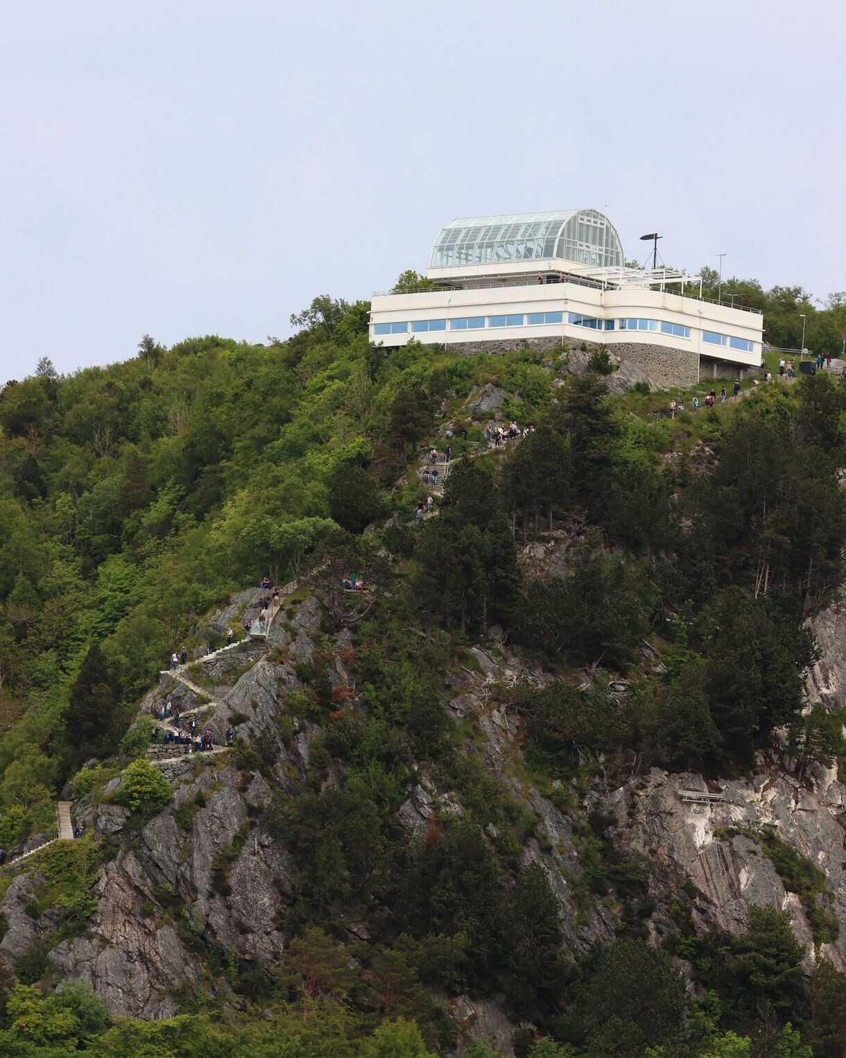 Modern glass-roofed Aksla Viewpoint building perched on a forested hillside above Ålesund, with a steep staircase trail visible below.