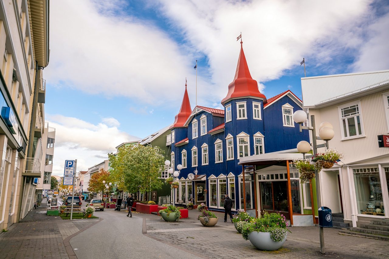 Street scene in Akureyri, Iceland, with a striking blue building topped by red spires, surrounded by shops, trees, flower planters and people walking under partly cloudy skies.