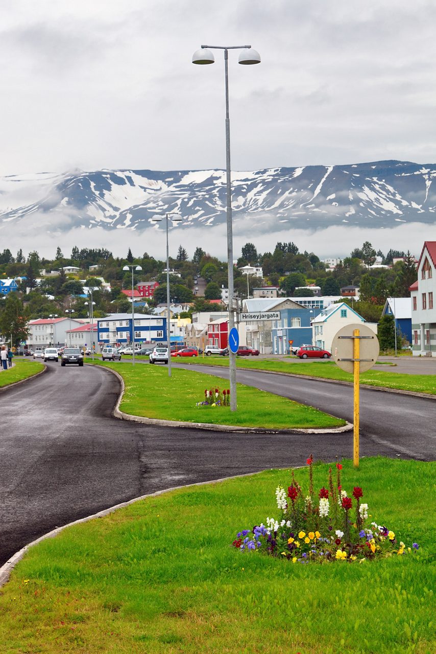 Street view of downtown Akureyri, Iceland, with colorful buildings, cars and flowerbeds along the road, set against a backdrop of snow-covered mountains and low-hanging clouds.