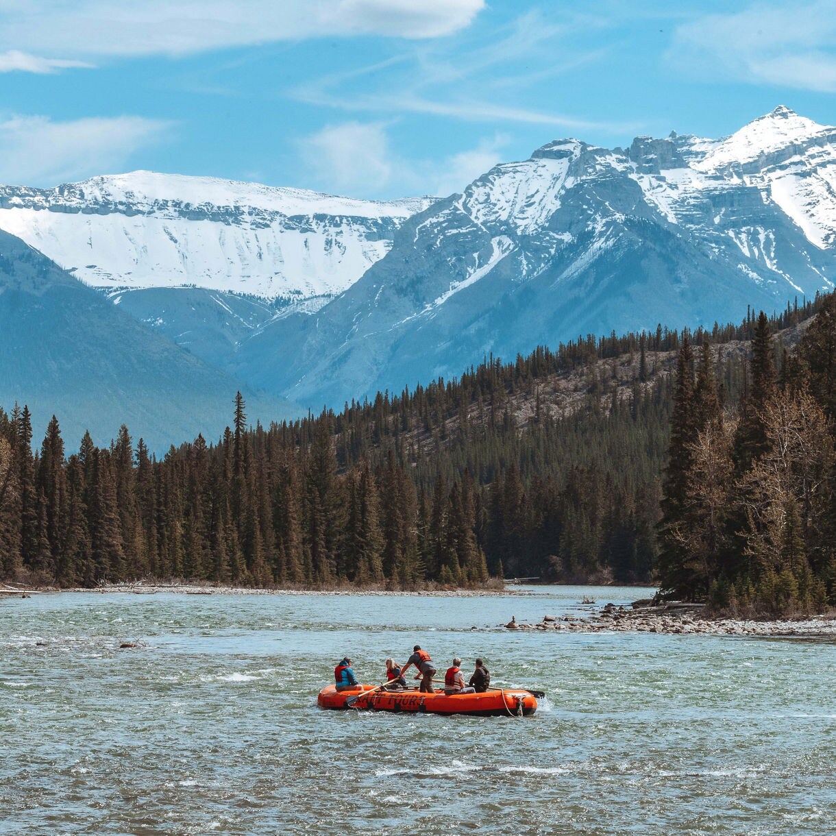 A group of rafters in an orange inflatable raft navigating a wide glacial river in Alaska, flanked by dense evergreen forests and dramatic snow-capped mountain peaks under a partly cloudy sky.