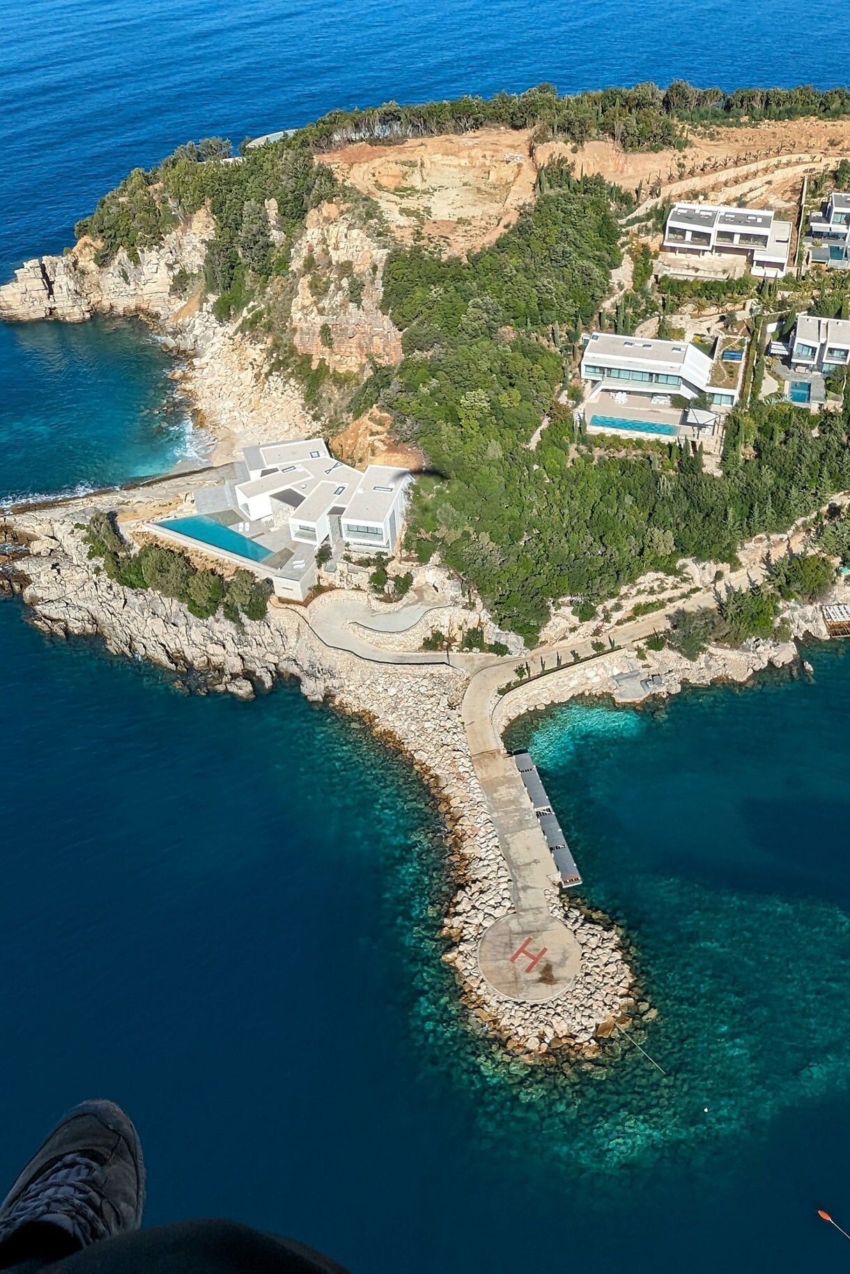 Aerial view of a rocky peninsula in southern Albania with a helipad at the tip, turquoise water surrounding it and modern villas with pools built into the lush hillside above.