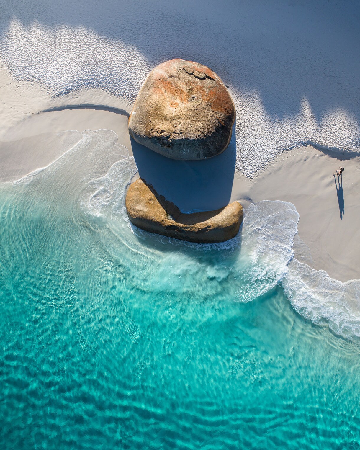 Aerial view of a bright turquoise shoreline with two large rounded boulders on white sand and a person walking nearby.