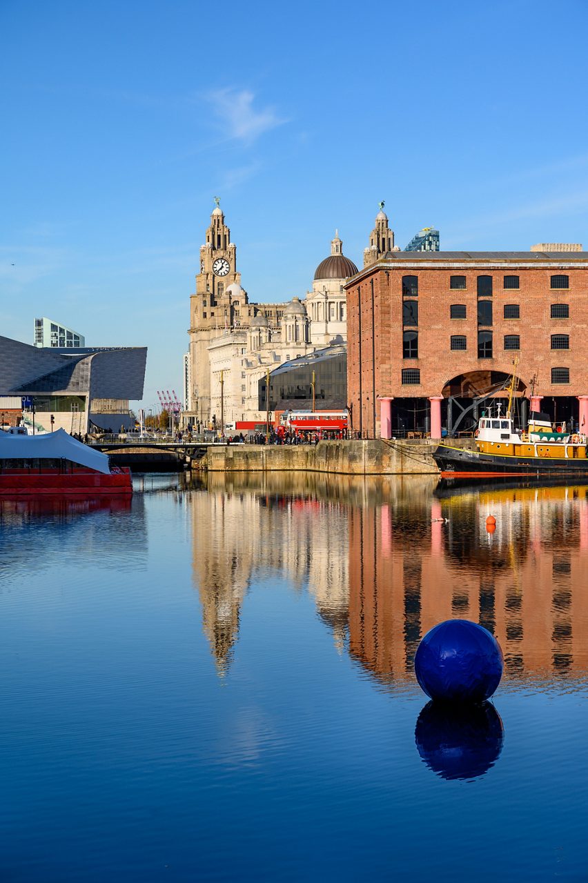 Waterfront view of Albert Dock in Liverpool with red-brick warehouses, boats and the Royal Liver Building in the background reflected in the calm water.
