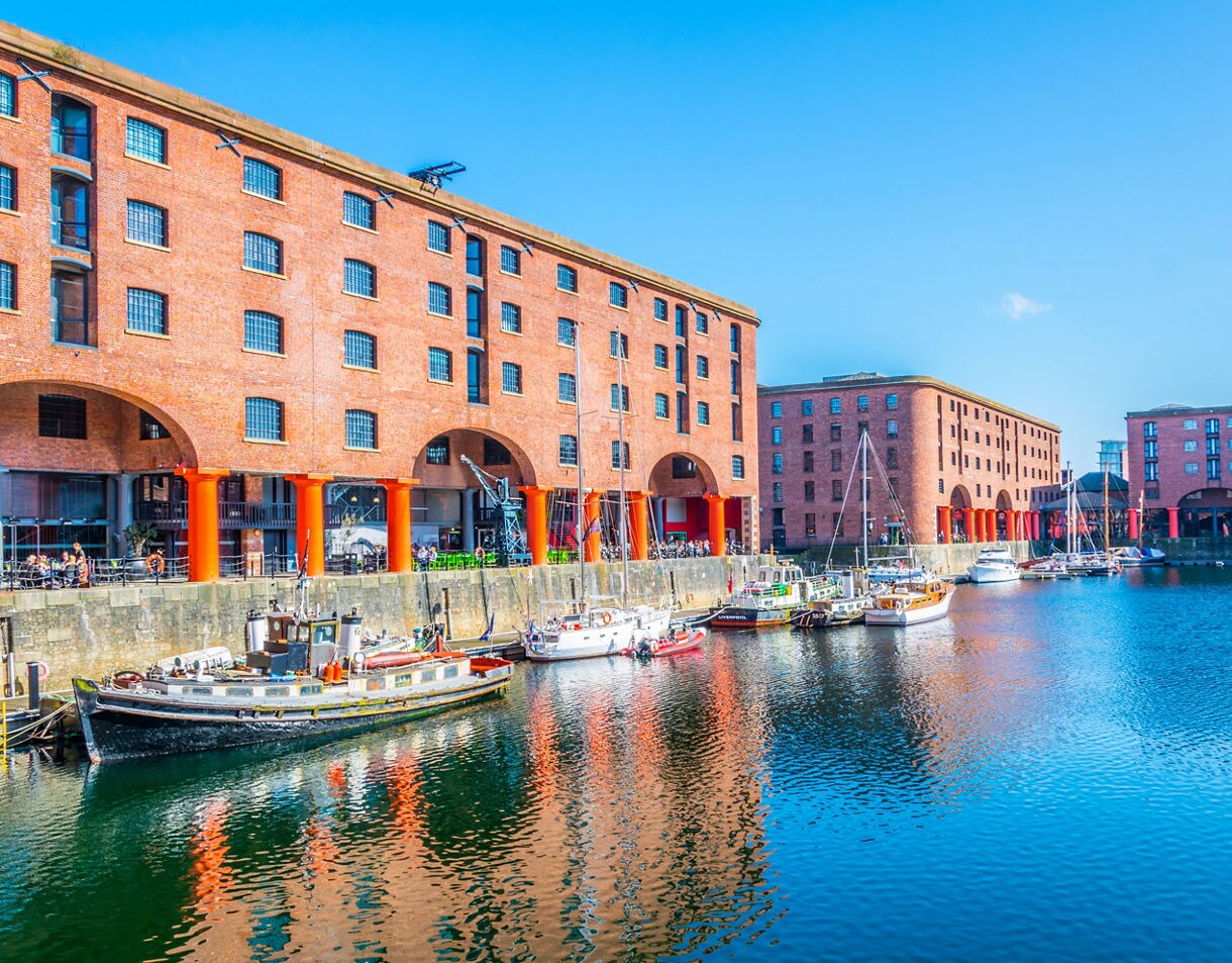 View of Albert Dock in Liverpool with moored boats and tall red-brick warehouses reflected in the calm water under a clear blue sky.