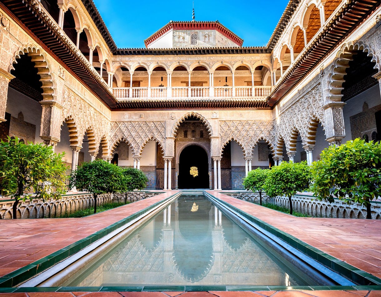 Courtyard of the Real Alcázar in Seville featuring ornate Moorish arches, a long reflecting pool and rows of orange trees beneath bright blue sky.