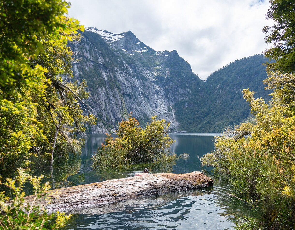 Clear lake bordered by dense green forest with steep rocky mountains rising in the background under soft, cloudy light.