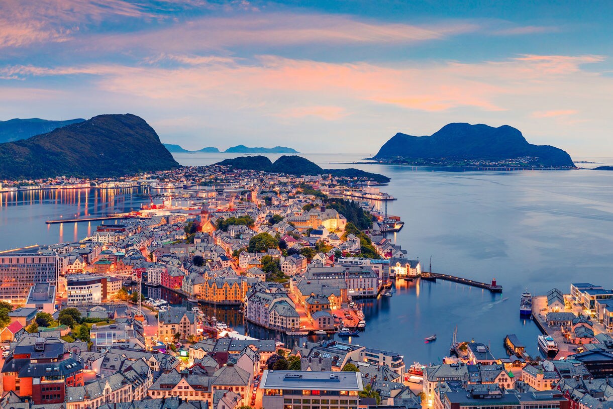 Aerial view of Ålesund, Norway at dusk with illuminated streets, colorful Art Nouveau buildings and surrounding fjords under a pastel sky.