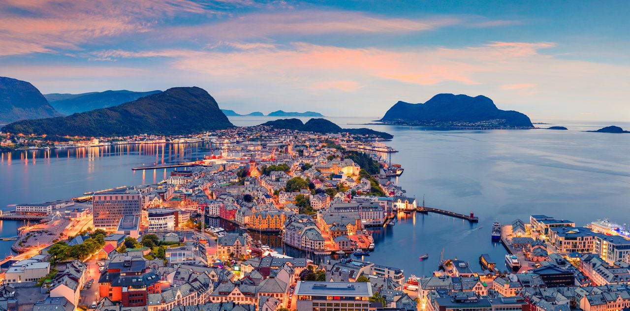 Aerial view of Ålesund, Norway at dusk with illuminated streets, colorful Art Nouveau buildings and surrounding fjords under a pastel sky.