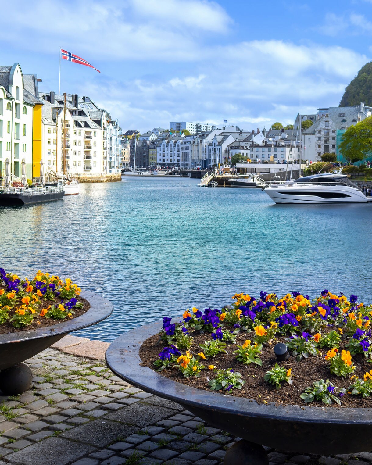 Colorful waterfront buildings and docked boats in Ålesund, Norway, with a Norwegian flag and bright flower planters in the foreground.