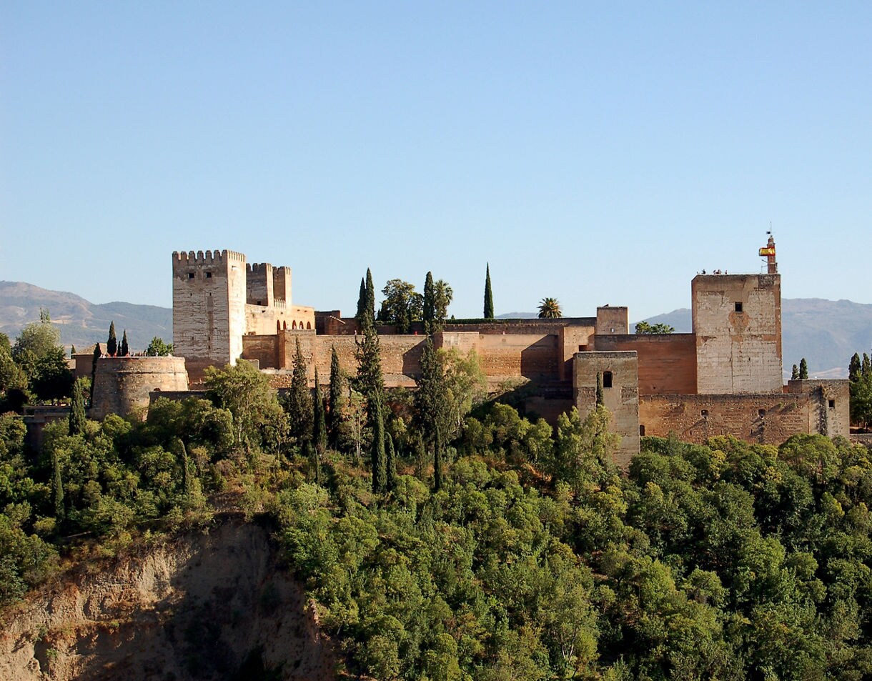 The Alhambra in Granada, Spain, with its ancient stone towers and walls surrounded by dense greenery under a clear sky.