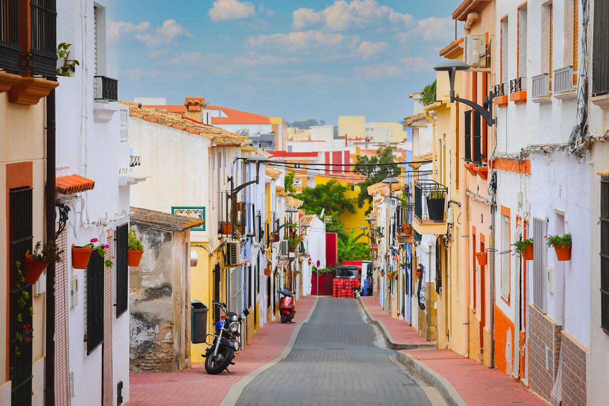 Narrow street in Alicante lined with brightly painted homes, potted flowers, balconies and parked scooters leading toward a sunlit backdrop of modern buildings.