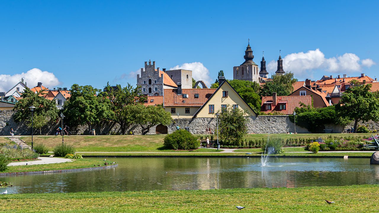 Almedalen Park in Visby, Gotland, Sweden, with a pond and fountain in the foreground, red-roofed houses and medieval stone towers rising in the background.