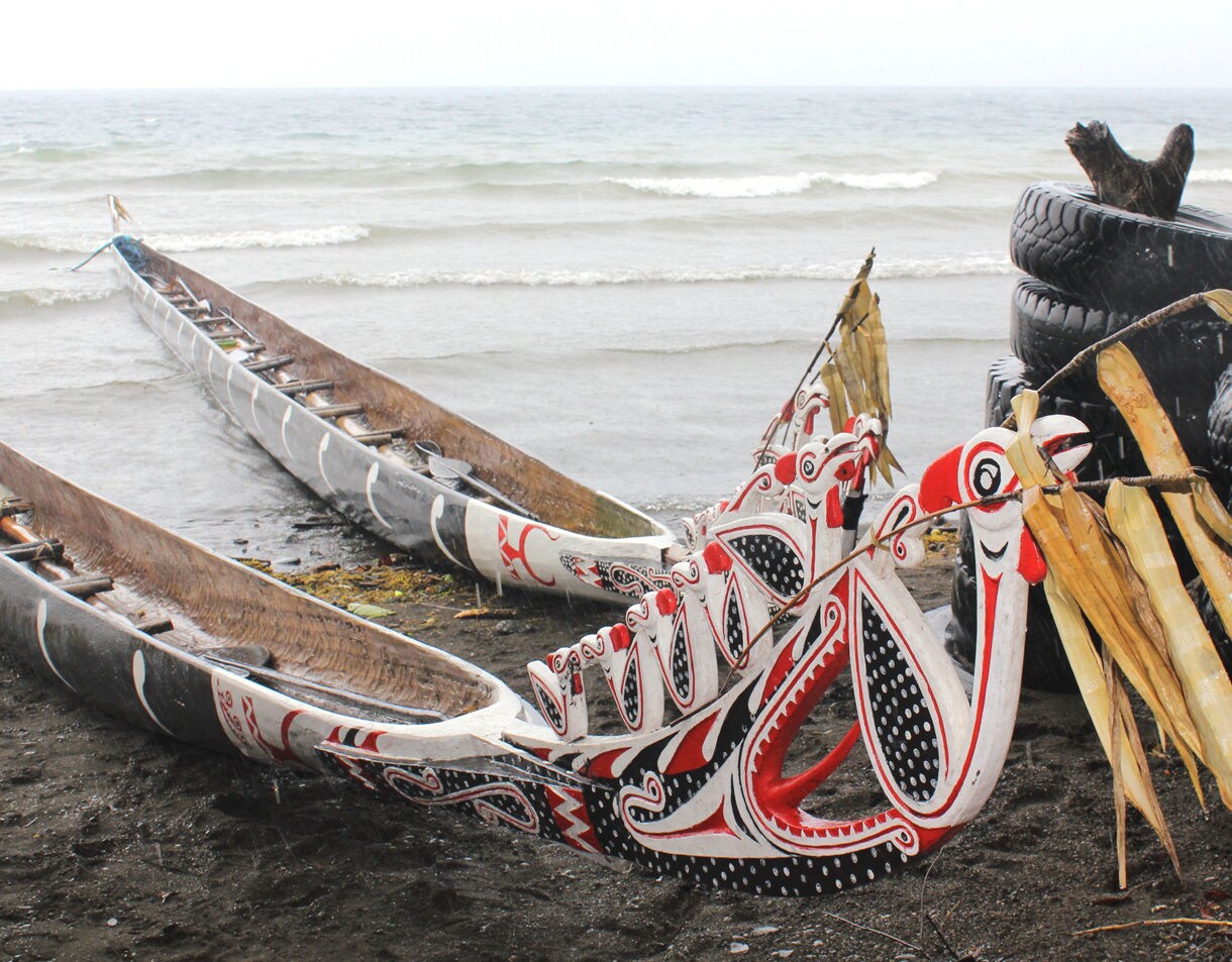 Long wooden canoes decorated with striking red, black and white painted designs, featuring carved bird-like prows, pulled onto a dark sandy beach with gentle waves rolling in behind them.