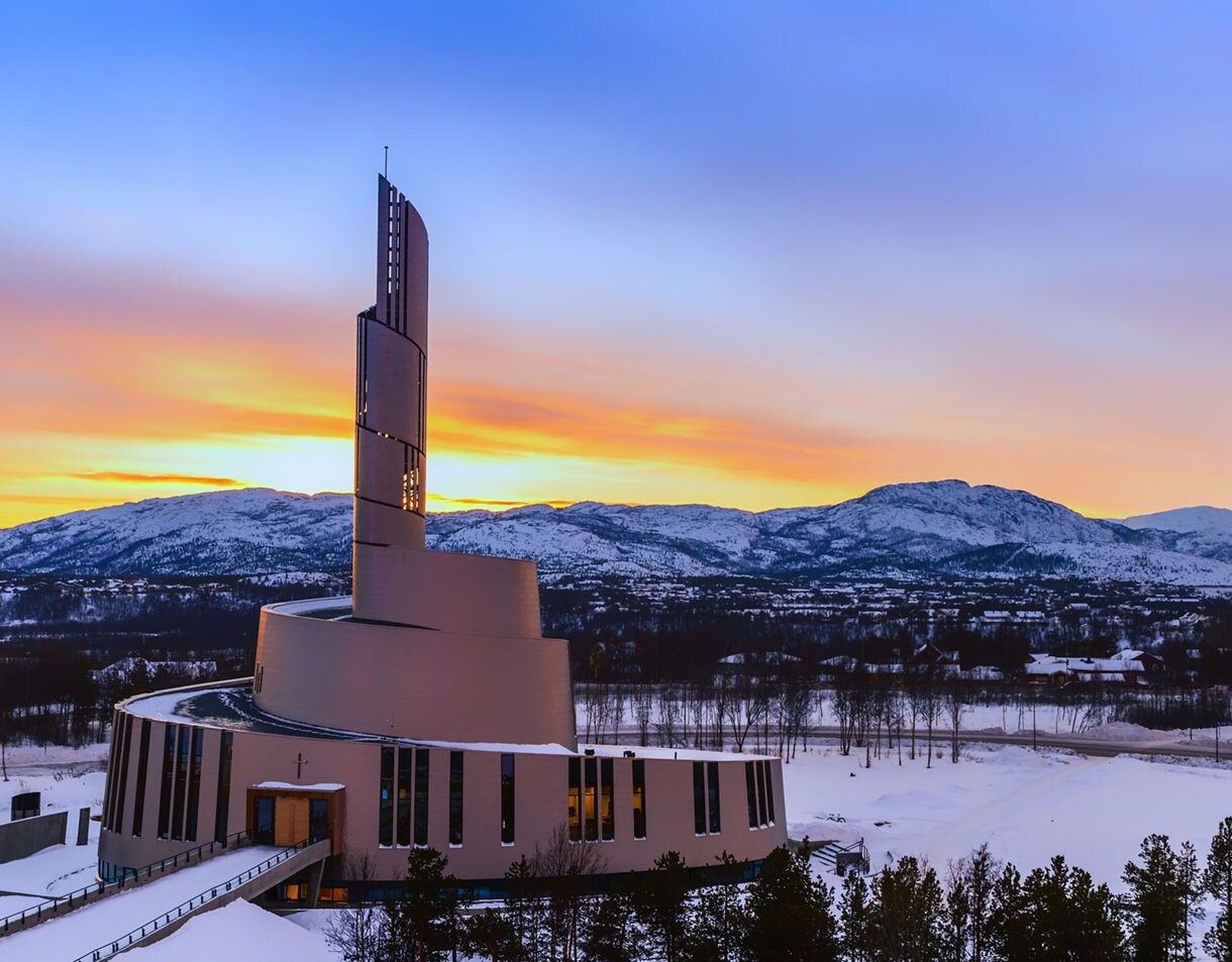 Modern spiraling Northern Lights Cathedral in Alta, Norway, set against snow-covered mountains and a vivid sunset sky.