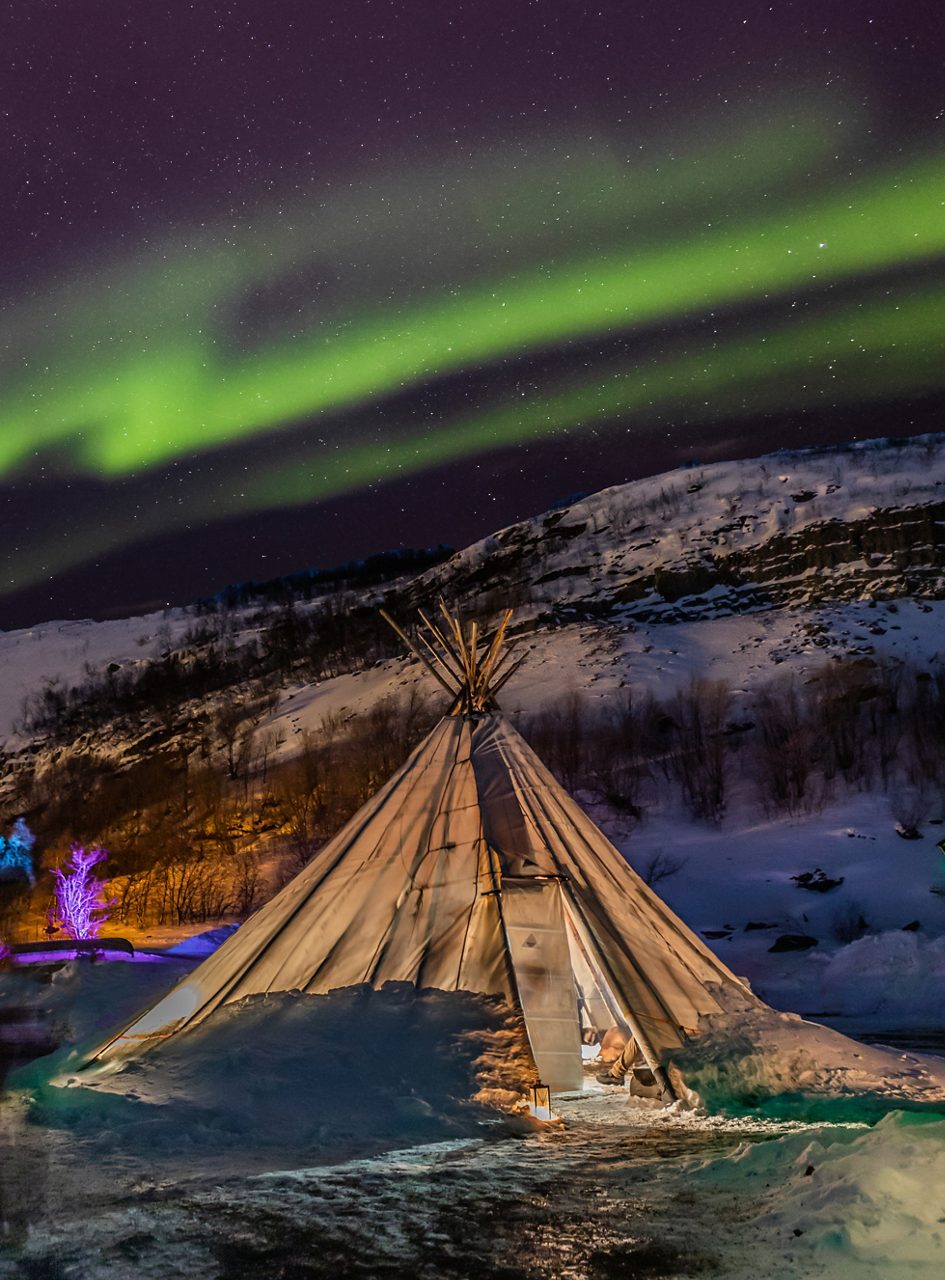 Traditional Sámi tent lit warmly at night under vivid green aurora borealis in a snowy valley.