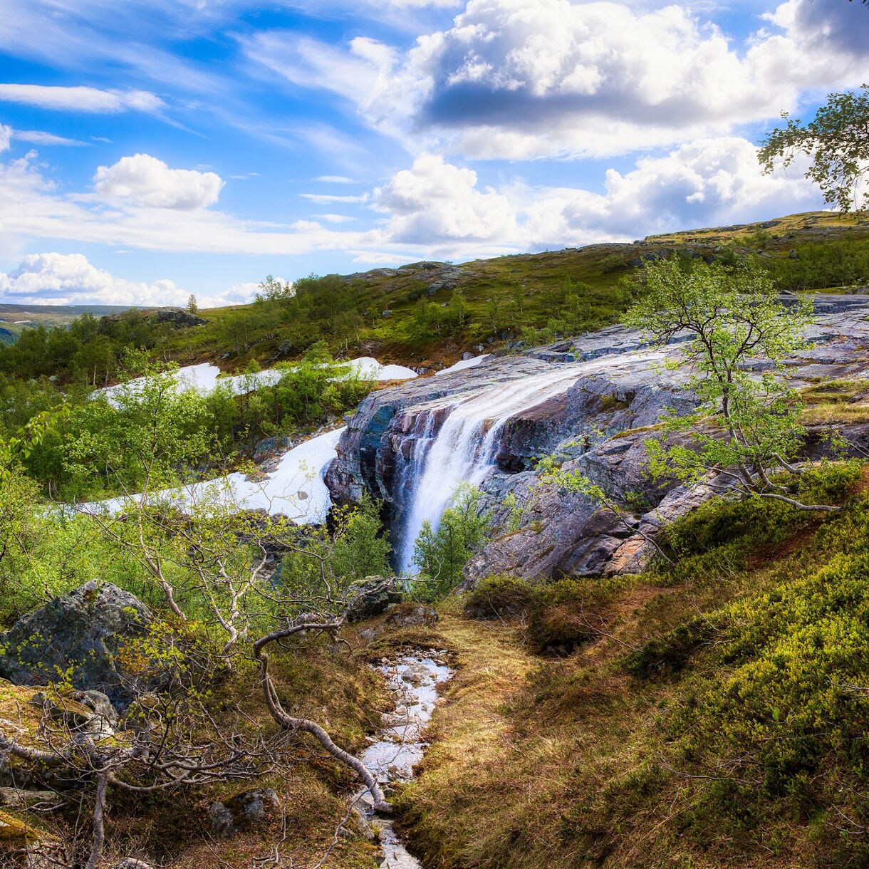 Cascading waterfall surrounded by green hills, rocks and patches of lingering snow in Alta, Norway.