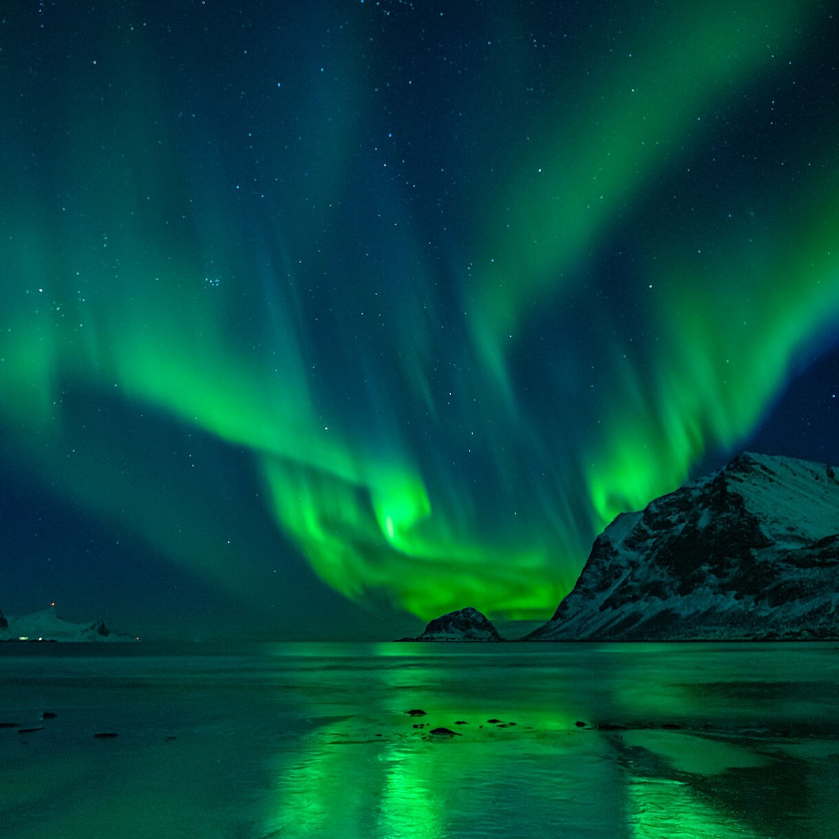 Bright green aurora borealis stretching over snowy mountains and reflecting on icy waters in Alta, Norway.