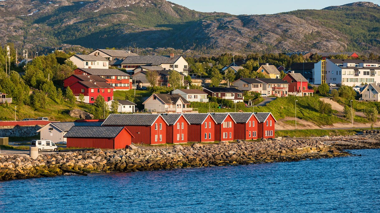 Coastal view of Alta with red wooden boathouses along the waterfront and houses scattered on green hills below rocky mountains.