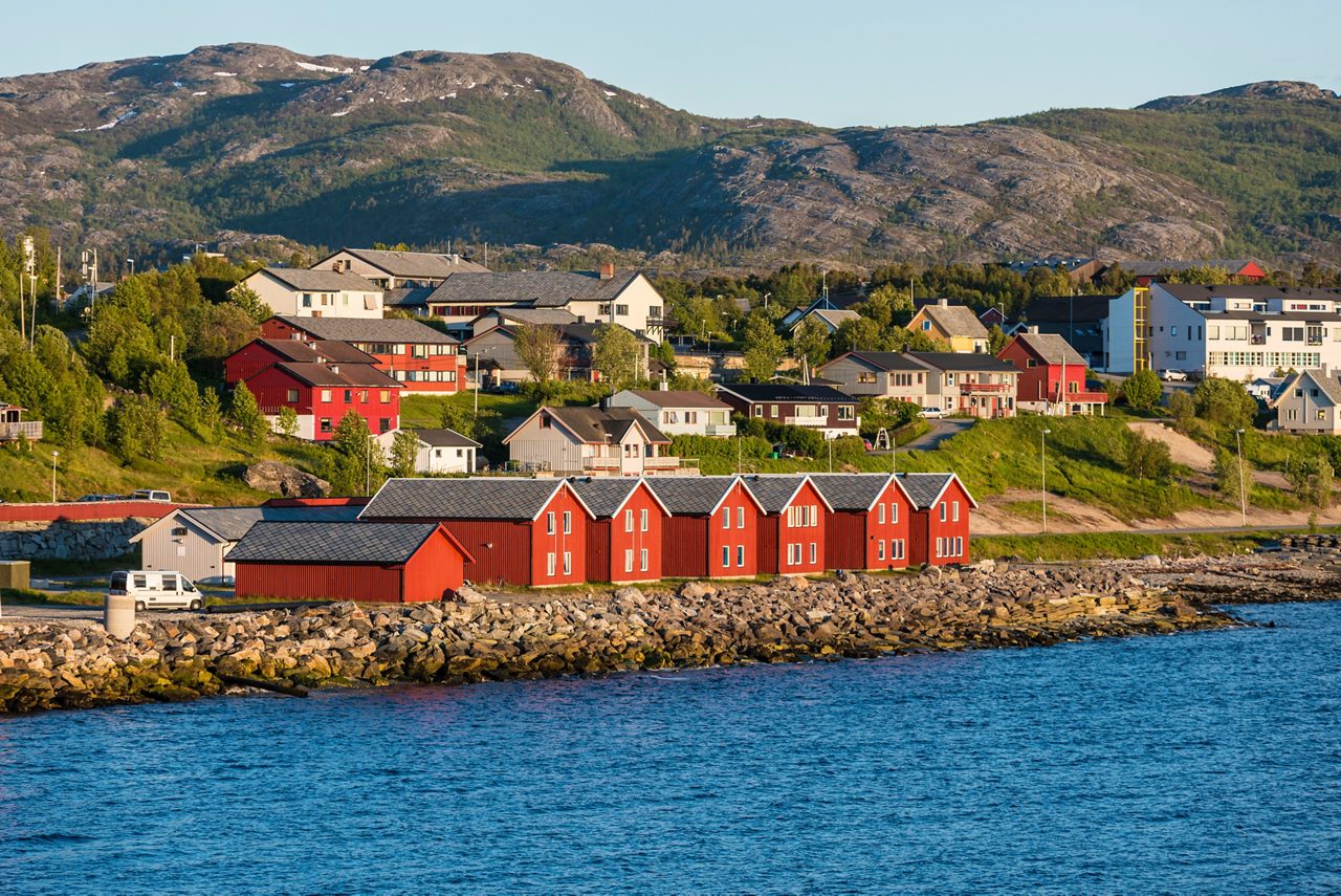 Coastal view of Alta with red wooden boathouses along the waterfront and houses scattered on green hills below rocky mountains.