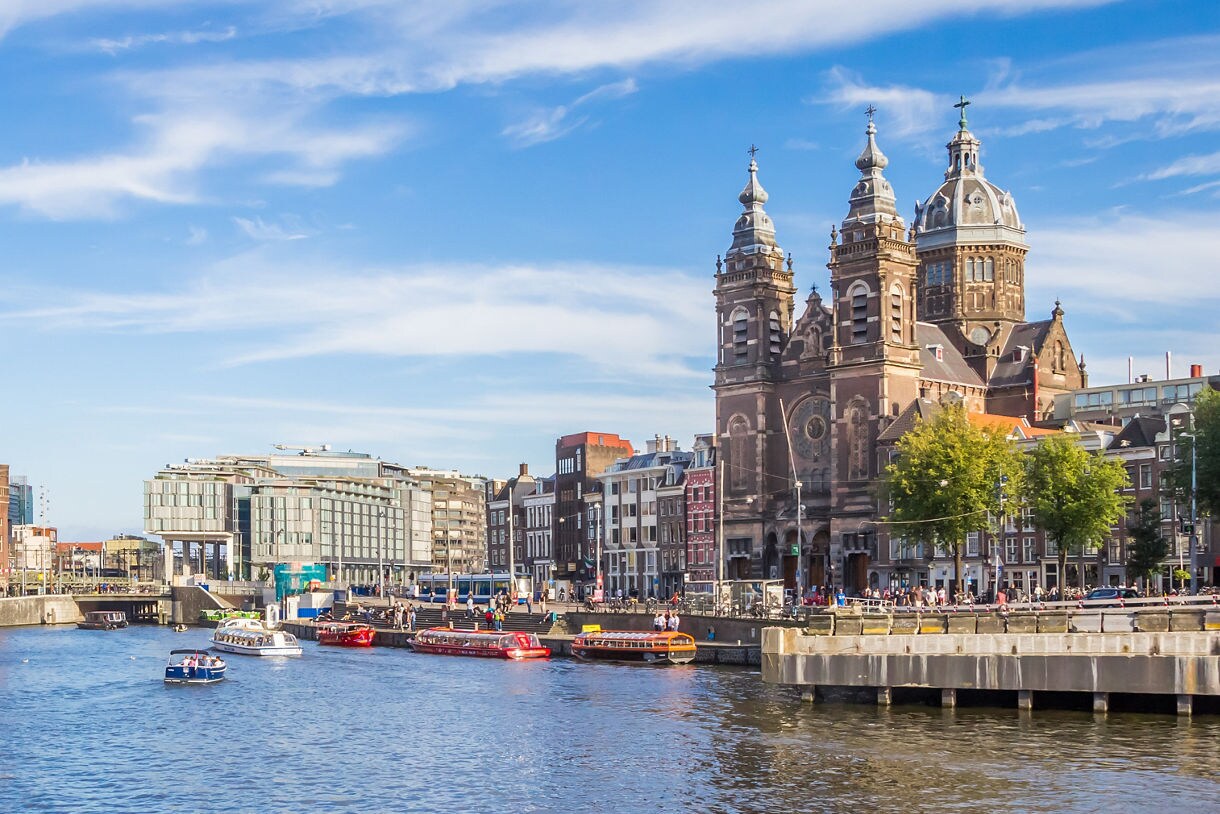 Scenic view of Amsterdam’s waterfront with canal boats on the water, historic Dutch buildings with red and brown facades and a large domed church rising behind them under a bright blue sky.