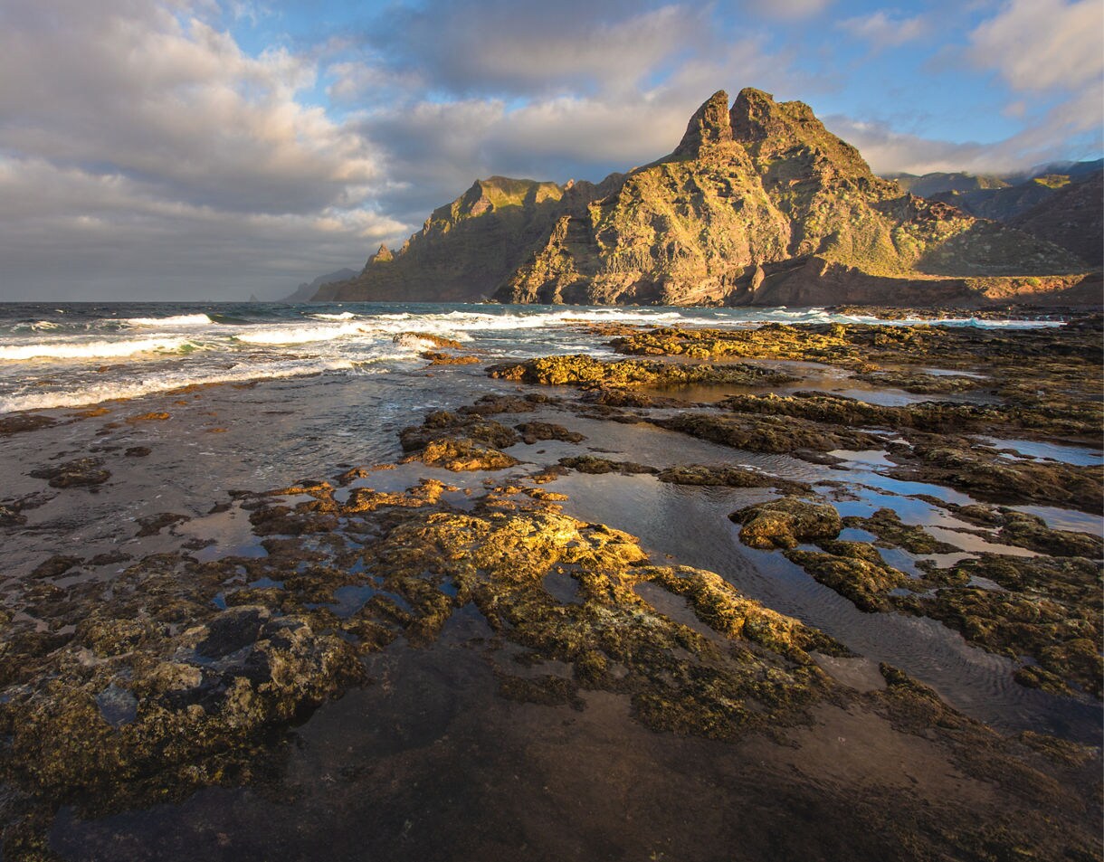 Rocky shoreline with tidal pools and waves crashing against the base of the sunlit Anaga Mountains under a cloudy sky in Tenerife.