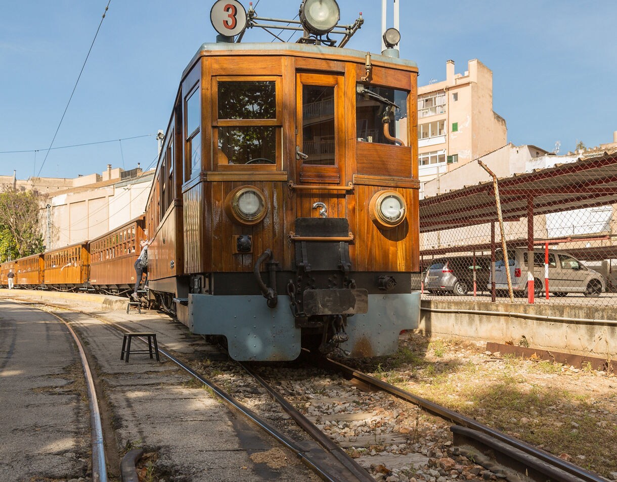 Close-up of a historic wooden train at the station in Palma, Mallorca, with passengers boarding and sunlight reflecting off the varnished carriages.