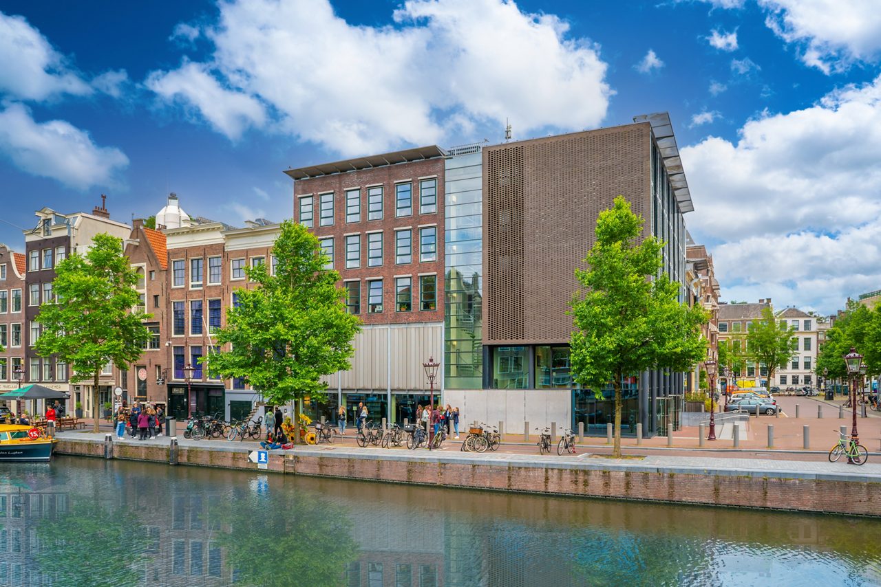 Exterior view of the Anne Frank House along an Amsterdam canal with modern brick and glass architecture, rows of traditional narrow houses, leafy trees and people walking and cycling beside the waterfront.