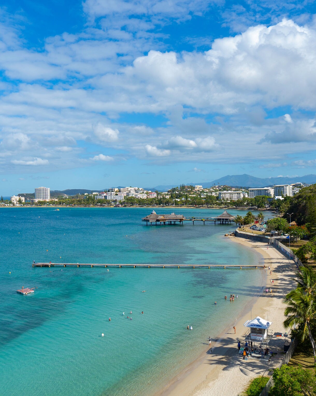 Elevated view of Anse Vata Beach in Nouméa with clear turquoise water, swimmers near the shore, a long beach lined with palm trees and a cityscape in the background under a bright blue sky.