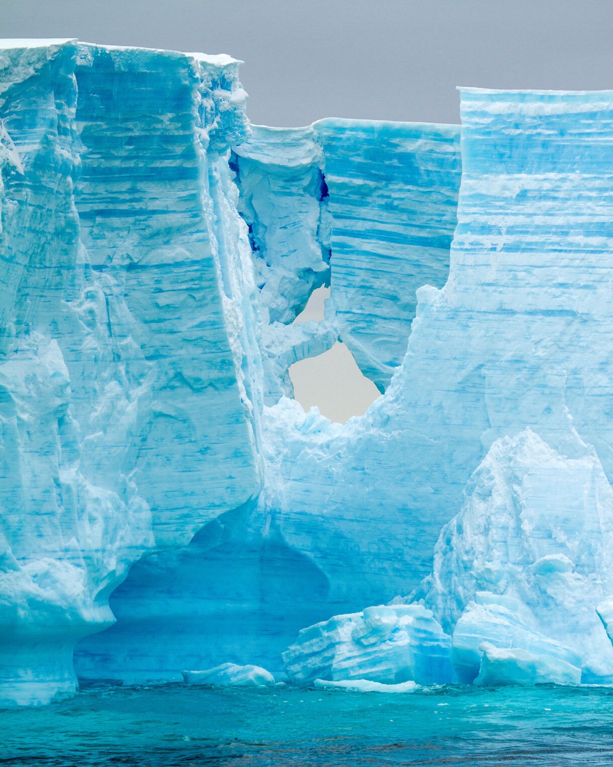 A massive blue glacier with deep ridges and a natural opening at its center rising above cold, dark Antarctic water.