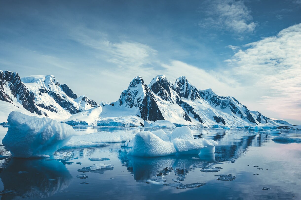 Snow-covered mountains reflected in calm Antarctic water with scattered blue icebergs floating across the scene.