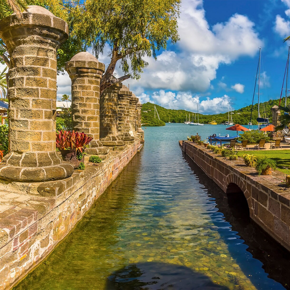 Stone pillars line a narrow waterway leading to a marina at English Harbour in Antigua, with palm trees, a green lawn and sailboats moored against hills in the background.