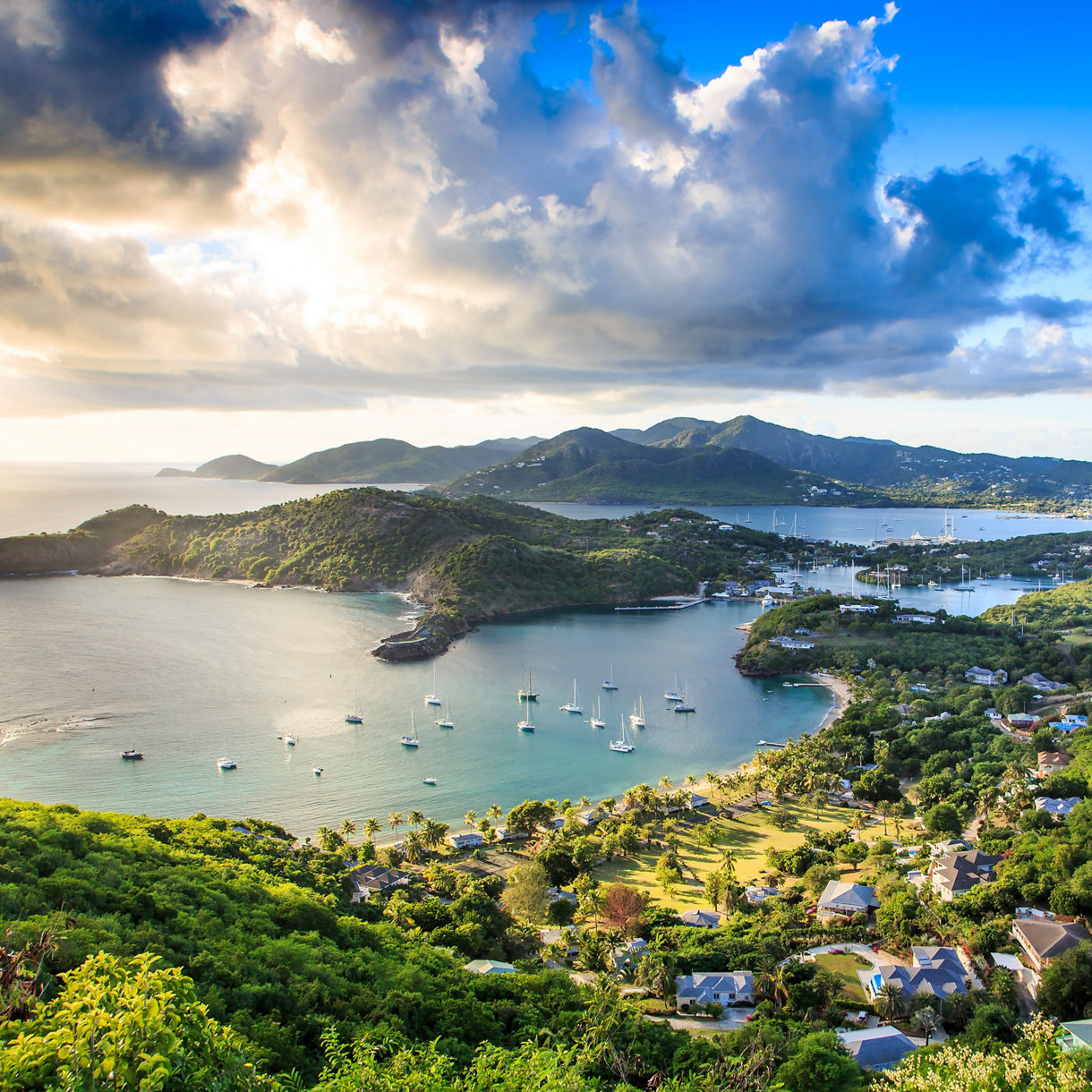 Aerial view of a lush green coastline in Antigua overlooking a harbor filled with sailboats, dramatic clouds gathering above the hills.