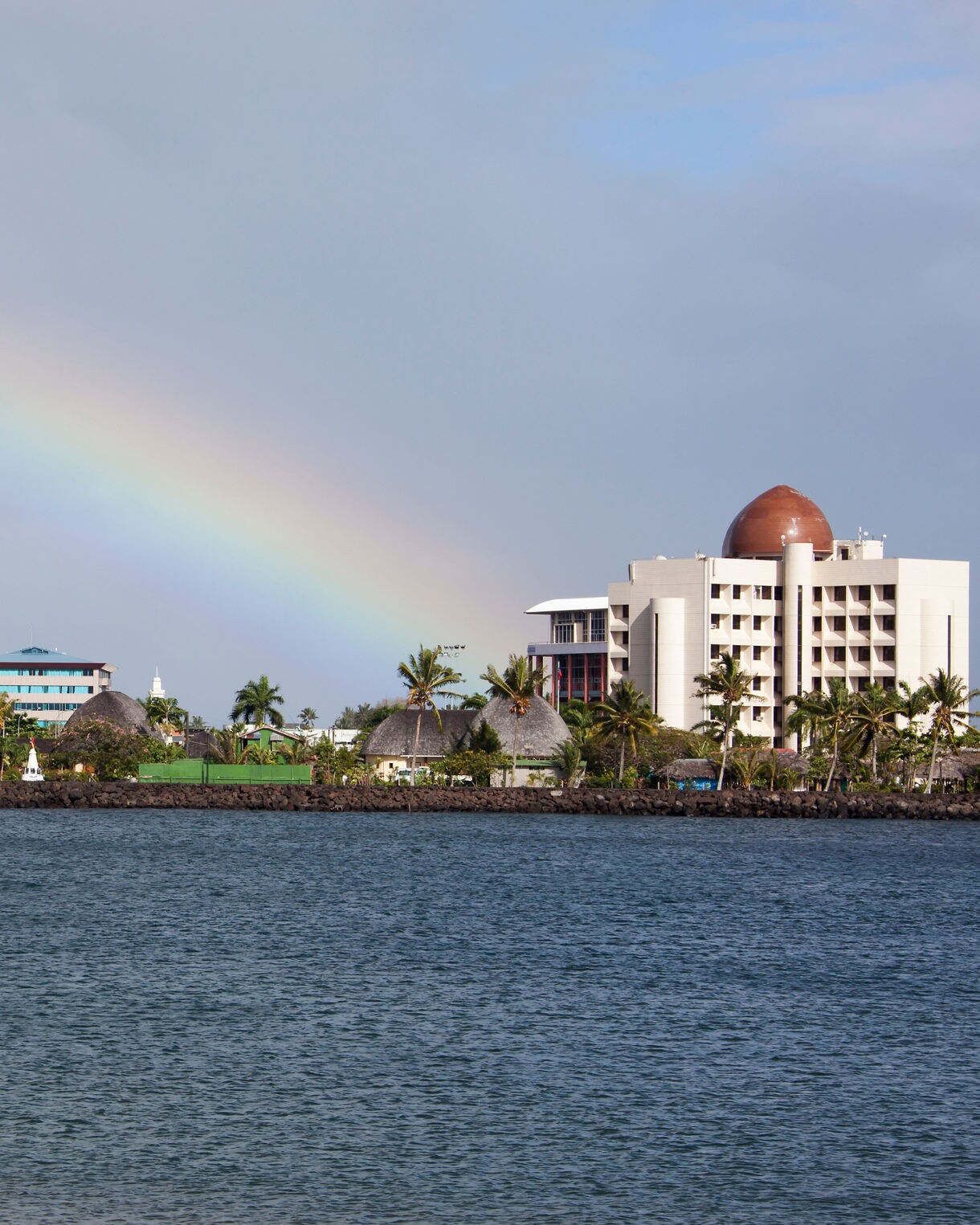 Coastal view of Apia, Samoa’s capital, featuring vibrant buildings, a domed government structure and a rainbow arching across the sky.