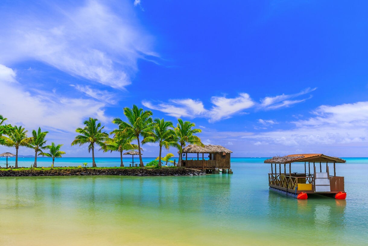 Palm trees and thatched huts overlooking a calm, turquoise lagoon in Apia, Samoa.