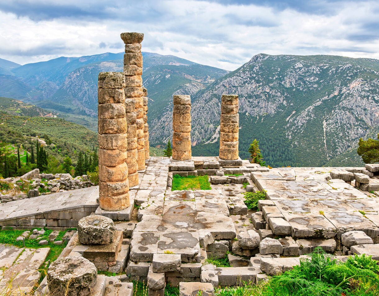 Stone columns and foundations of Apollo’s Sanctuary in Delphi, overlooking rugged green mountains and valleys under a cloudy sky.