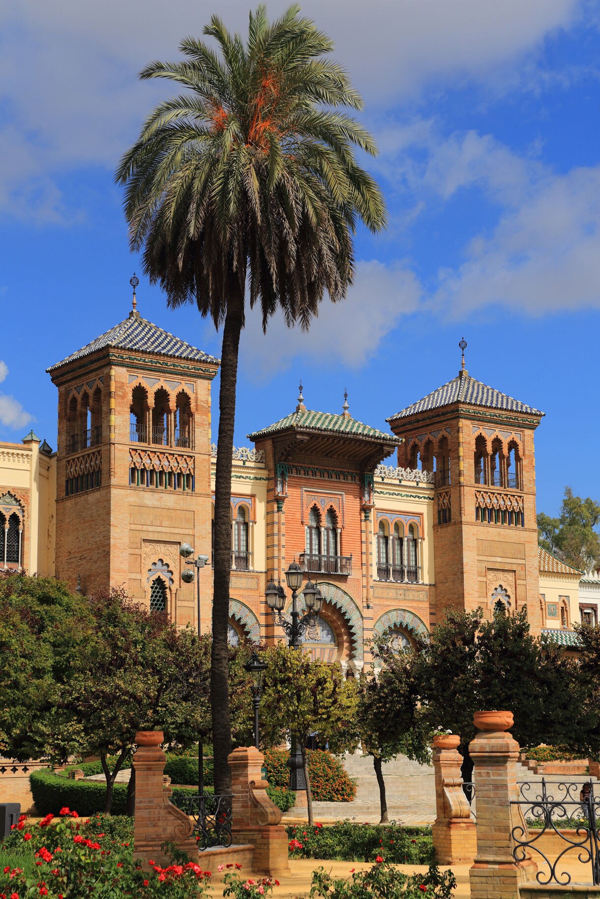 Mudéjar Pavilion in Seville with ornate arches, patterned brickwork and tiled roofs, framed by palm trees and garden greenery under a bright blue sky.