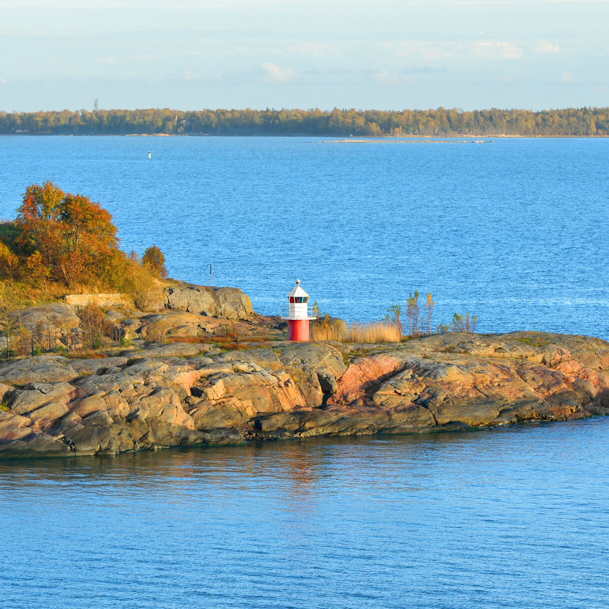 Small red-and-white lighthouse on a rocky island in the Finnish archipelago, surrounded by calm blue waters with a sailboat in the distance.