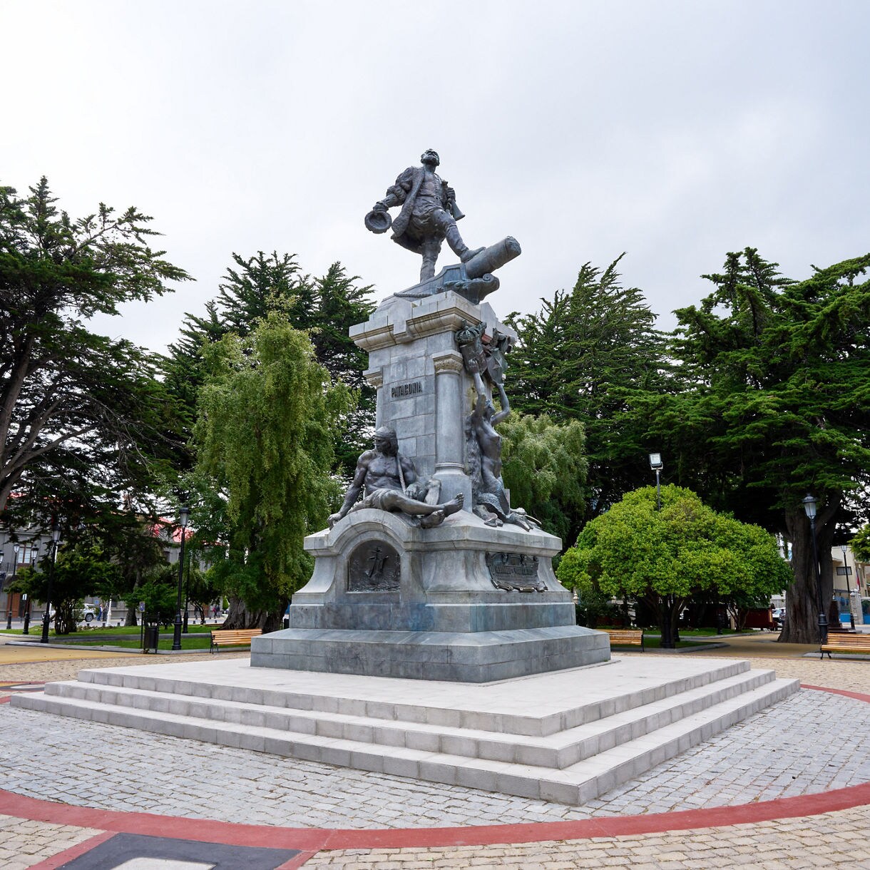 Monument to Ferdinand Magellan at the center of Plaza Muñoz Gamero, surrounded by tall trees, benches and a circular paved plaza.