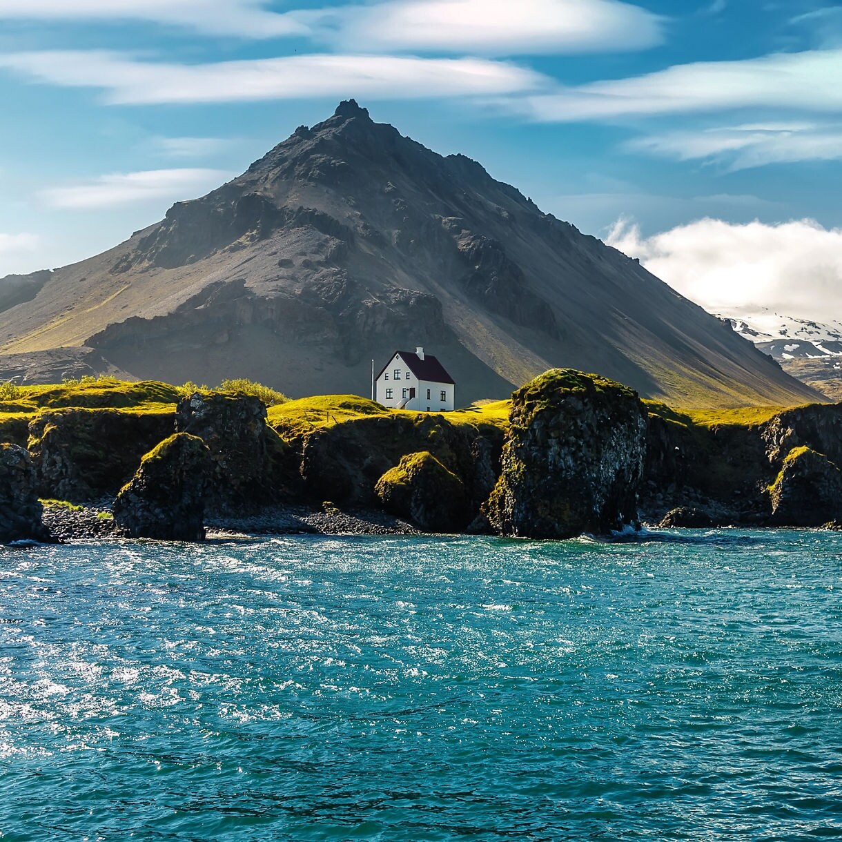 A small white house with a red roof perched on grassy cliffs above deep blue coastal water, with rugged volcanic mountains rising in the background under a bright sky.