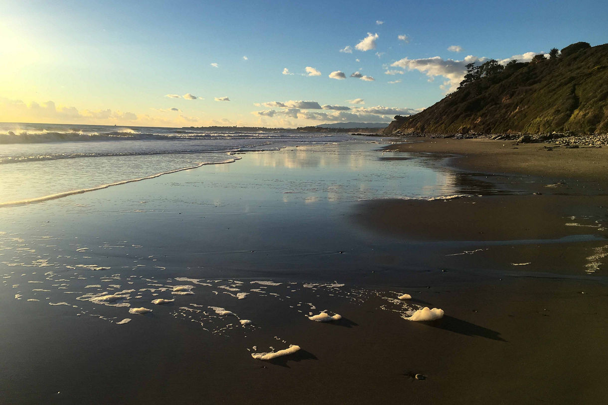 A serene beach scene at Arroyo Burro Beach during late afternoon, with gentle waves washing onto the wet sand, reflecting the golden light of the setting sun.
