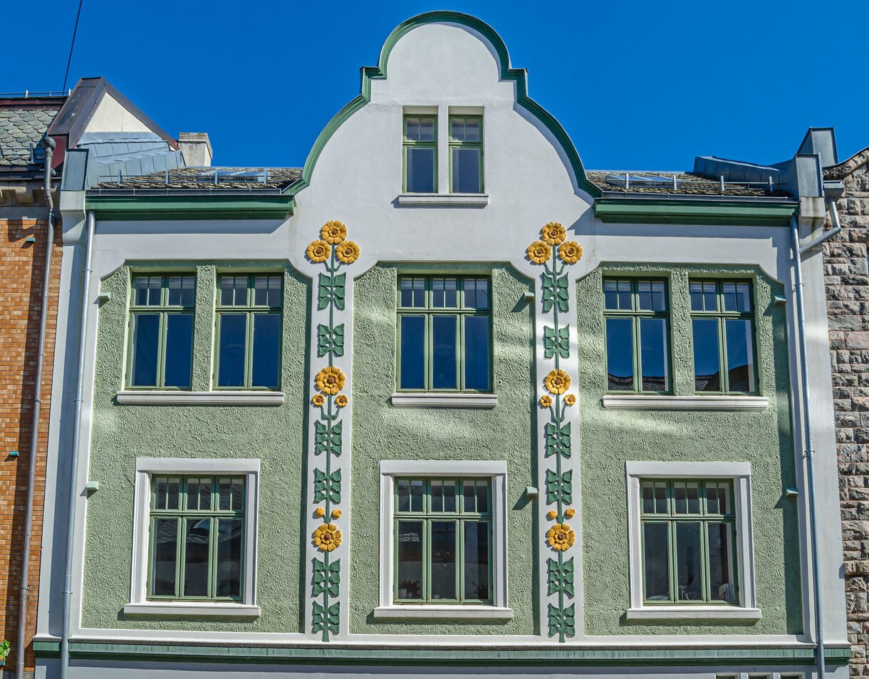Decorative Art Nouveau building in Ålesund with green walls, yellow floral motifs and symmetrical windows under a bright blue sky.