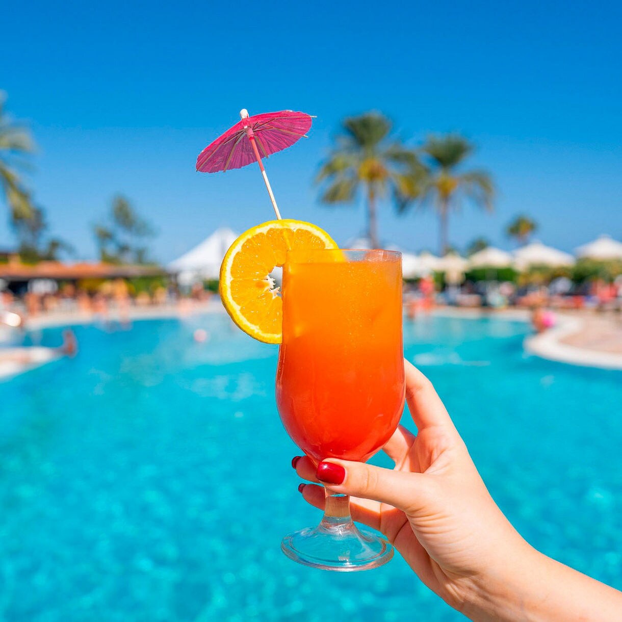 Hand holding an orange-red Aruba Ariba cocktail garnished with an orange slice and paper umbrella in front of a bright blue resort pool with palm trees and sunbathers in the background.
