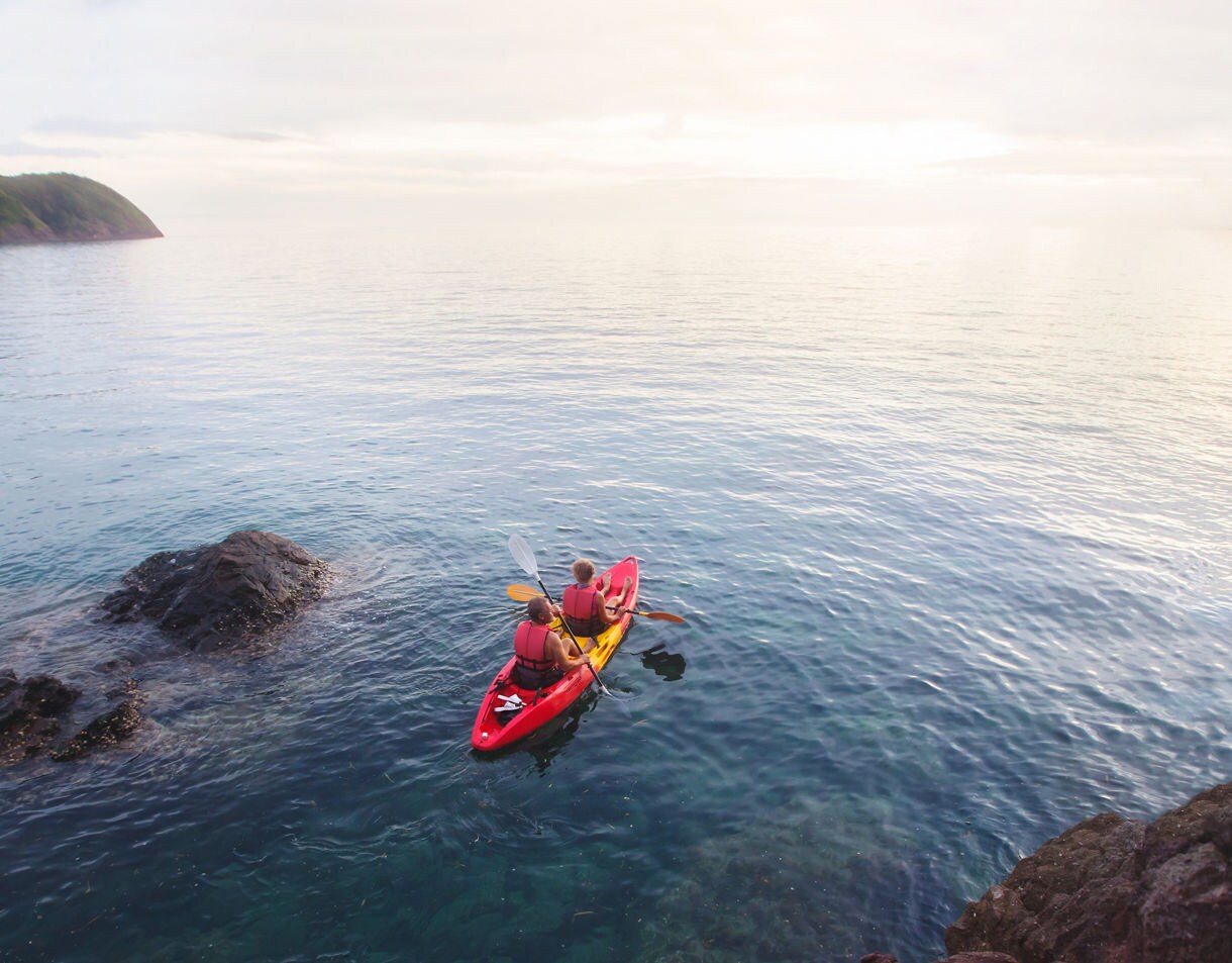 Two people in a red and yellow tandem kayak paddling over clear, tranquil water near rocky outcrops with distant tree-covered hills under a bright, hazy sky.